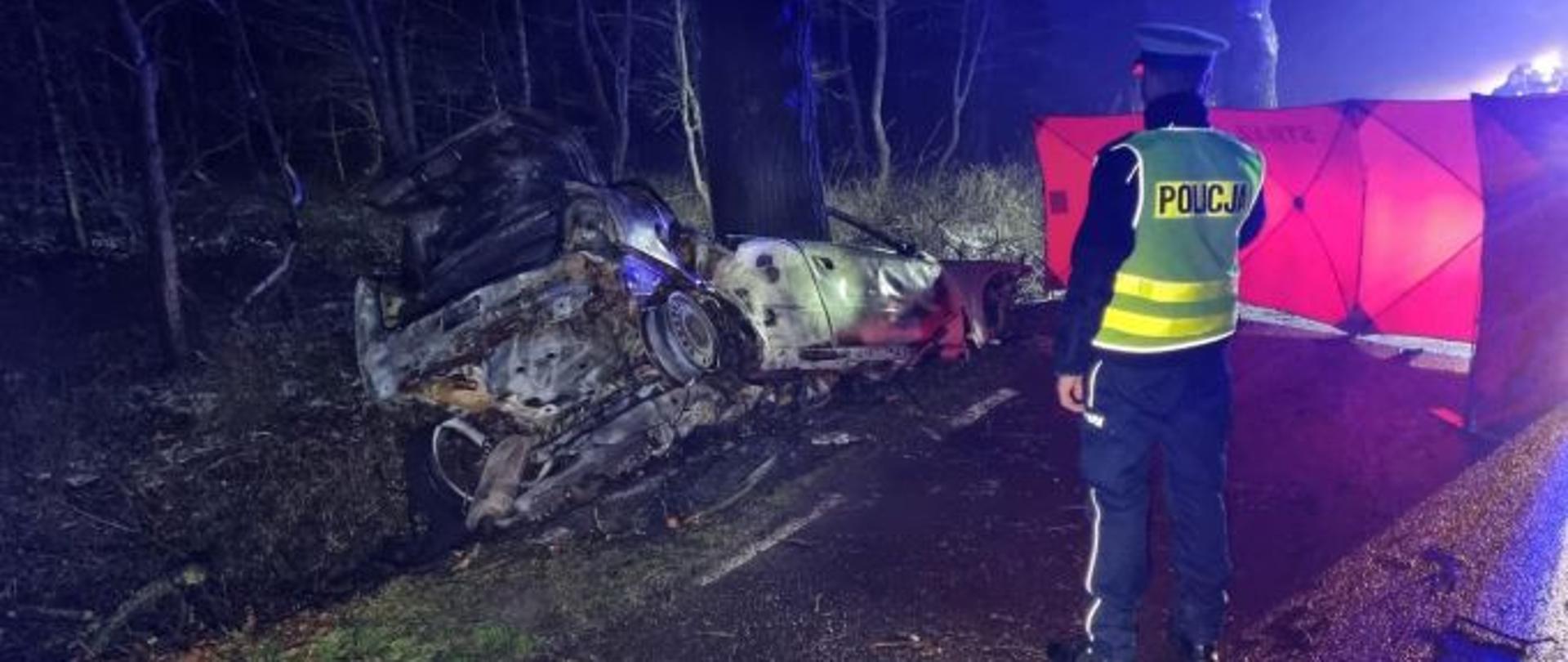 Pora nocna. Zdjęcie przedstawia spalony pojazd leżący na poboczy drogi w okolicy drzewa, z tyłu kadru las. Z prawej strony stoi policjant obok niego znajduje się czerwony parawan osłaniający miejsce zdarzenia.