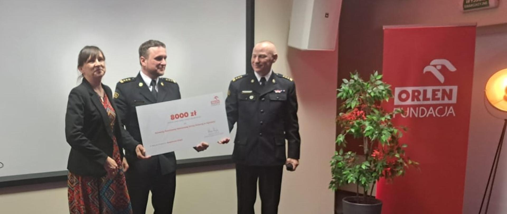 Na tle białego ekrany stoi dwóch mężczyzn i kobieta. Mężczyźni trzymają wielką kartkę.