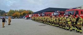 Na placu przed obiektem jednostki ochrony przeciwpożarowej odbywa się zbiórka strażaków. W szeregu ustawieni są funkcjonariusze PSP oraz druhowie OSP w ubraniach specjalnych, a za nimi zaparkowane są samochody ratowniczo-gaśnicze. Przed zebranymi występują osoby prowadzące odprawę. Zdjęcie przedstawia przygotowanie sił i środków ćwiczeń.