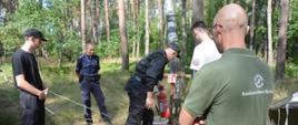 Zdjęcie przedstawia funkcjonariuszy Komendy Powiatowej Państwowej Straży Pożarnej w Krośnie Odrzańskim, funkcjonariusza Komendy Powiatowej Policji w Krośnie Odrzańskim, przedstawiciela Nadleśnictwa Bytnica oraz Komendanta Obozu Harcerskiego „Struga” podczas sprawdzania punktu przeciwpożarowego na terenie obozu. Zdjęcie wykonane zostało na tle terenów leśnych w Obozie Harcerskim „Struga”.