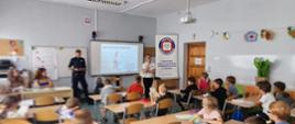 Policjantka oraz kobieta w kremowej bluzce stoją przed grupą siedzących na krzesłach dzieci. Widoczny jest roll-up z logo Państwowej Inspekcji Sanitarnej