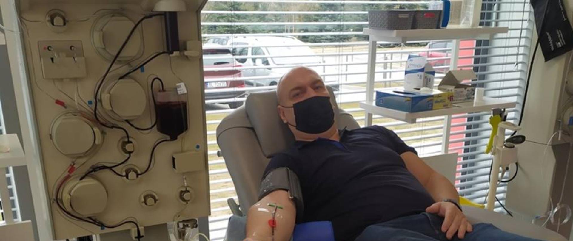 Zdjęcie przedstawia pracownika Komendy Powiatowej Państwowej Straży Pożarnej w Zambrowie w ubraniu cywilnym podczas oddawania osocza. W tle widoczna jest aparatura służąca do pobierania osocza.