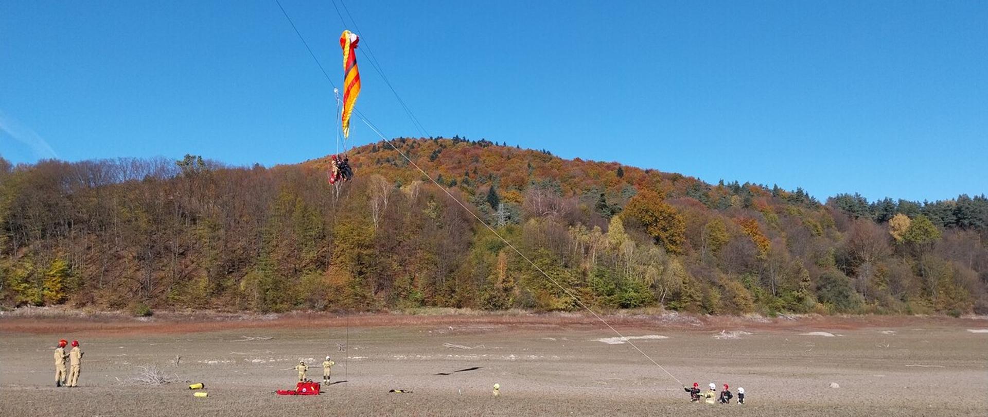 Na zdjęciu widoczny na tle lasu paralotniarz który zawisł na linii średniego napięcia w miejscowości Uście Gorlickie poniżej strażacy z Państwowej Straży Pożarnej i Ochotniczych Straży Pożarnych ubrani w ubrania służbowe i specjalne koloru piaskowego.