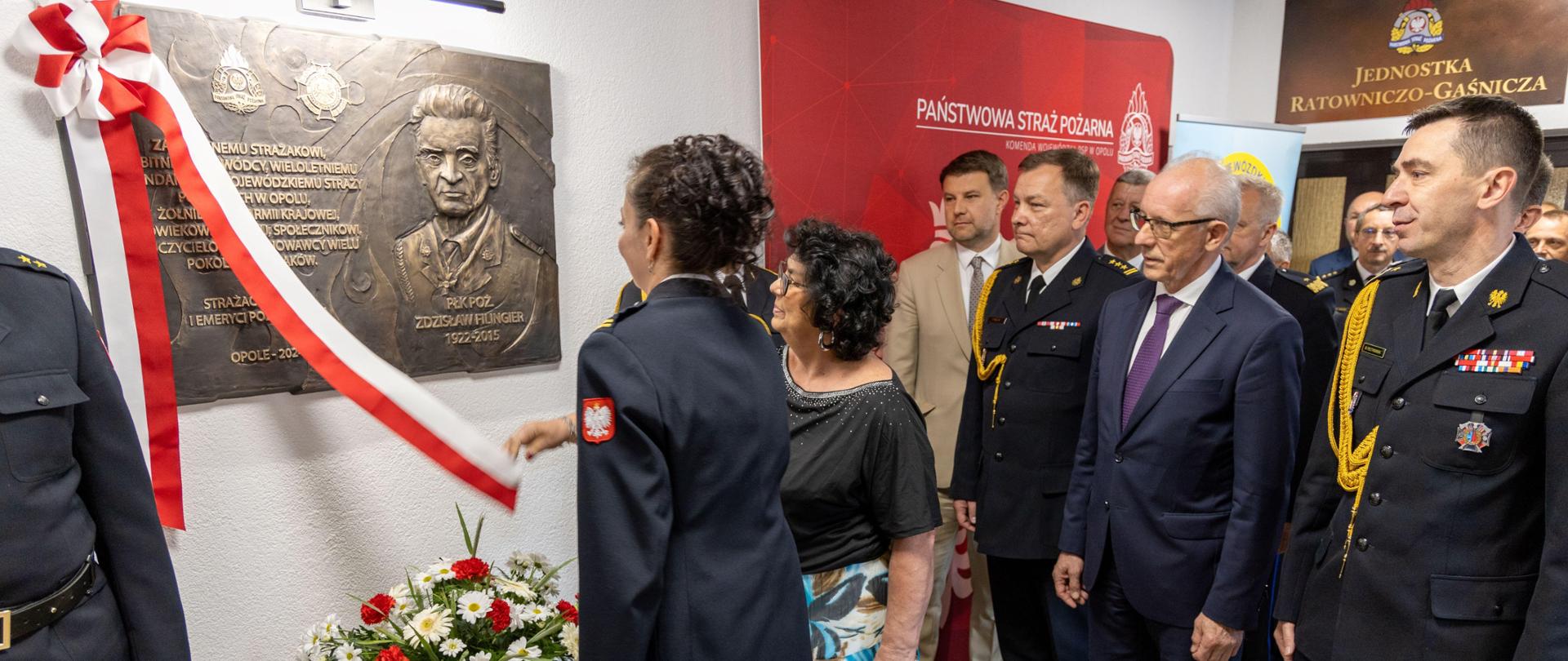 Dwie kobiety odsłaniają pamiątkową tablicę płk. poż. Zdzisława Filingier. Kobiety odsłaniające tablicę to córka i wnuczka płk. poż. Zdzisława Filingiera. Obok stoi Komendant Główny PSP i podsekretarz stanu w Ministerstwie Spraw Wewnętrznych i Administracji, a także opolski komendant wojewódzki PSP.