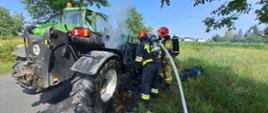 Zdjęcie przedstawia palącą się ładowarkę teleskopową koloru zielonego oraz strażaków PSP podczas gaszenia pożaru. W tle pojazd pożarniczy koloru czerwonego.
