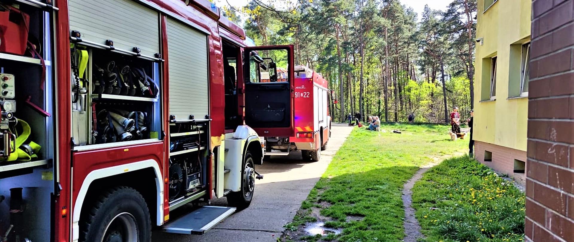 Dwa samochody pożarnicze po lewej stronie stoją na drodze dojazdowej do bloku mieszkalnego. Po prawej blok mieszkalny. W tle na trawniku osoby ewakuowane i las.
