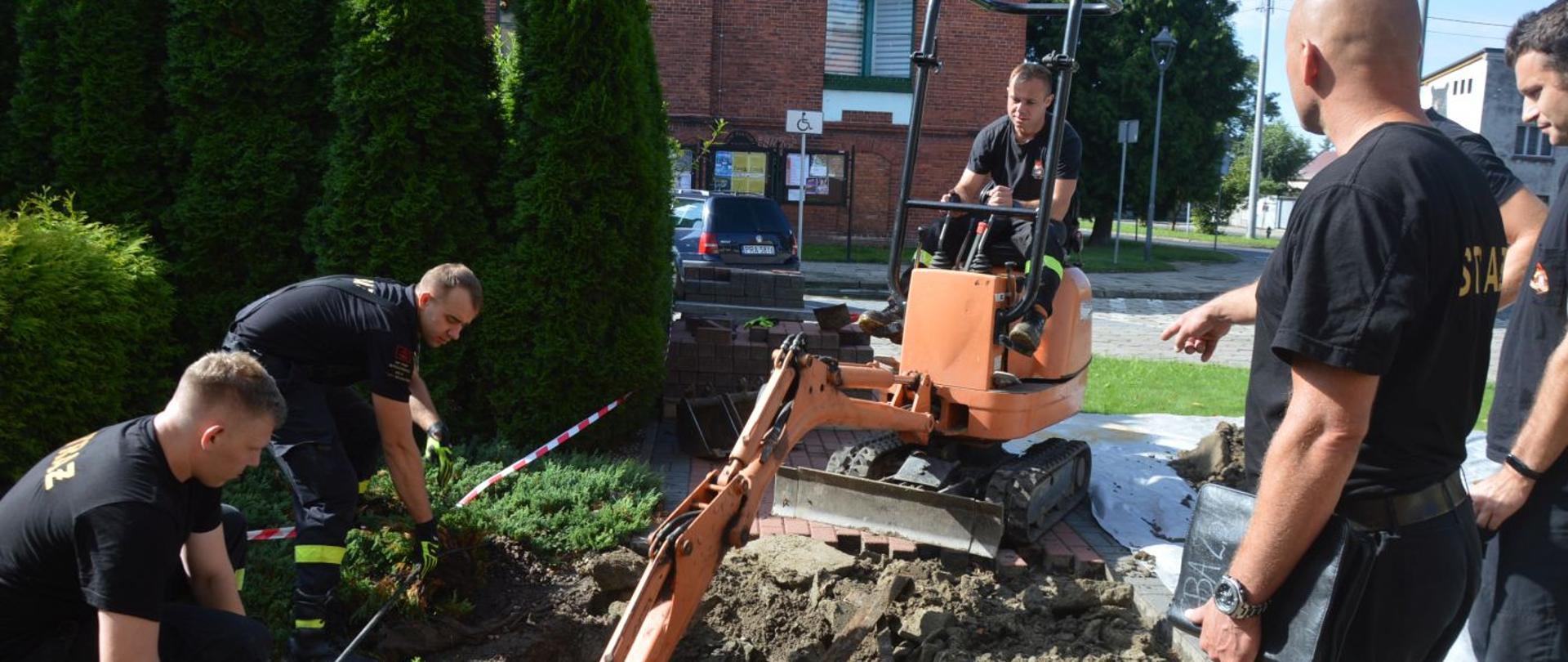 Remont chodnika. Teren przed budynkiem Komendy Powiatowej PSP w Rawiczu. Pięciu strażaków wykonuje prace remontowe chodnika. Jeden z nich operuje minikoparką.W tle tuje oraz fragment budynku domu kultury.