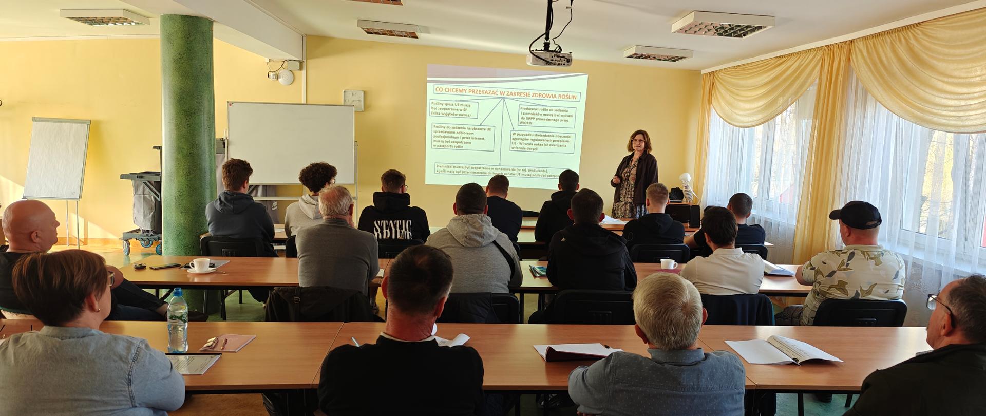 Uczestnicy spotkania siedzą przy długich stołach w sali szkoleniowej i słuchają prelegentki stojącej przy ekranie projekcyjnym. Na ekranie wyświetlana jest prezentacja, a prowadząca omawia jej treść. Uczestnicy mają przed sobą notatniki i materiały szkoleniowe. Sala jest dobrze oświetlona, z dużymi oknami zasłoniętymi jasnymi zasłonami.