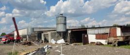 Zdjęcia w ciągu dnia. Widać teren działań ratowniczo-gaśniczych, samochód specjalny w kolorze czerwonym. W tle zabudowania gospodarcze oraz silos.