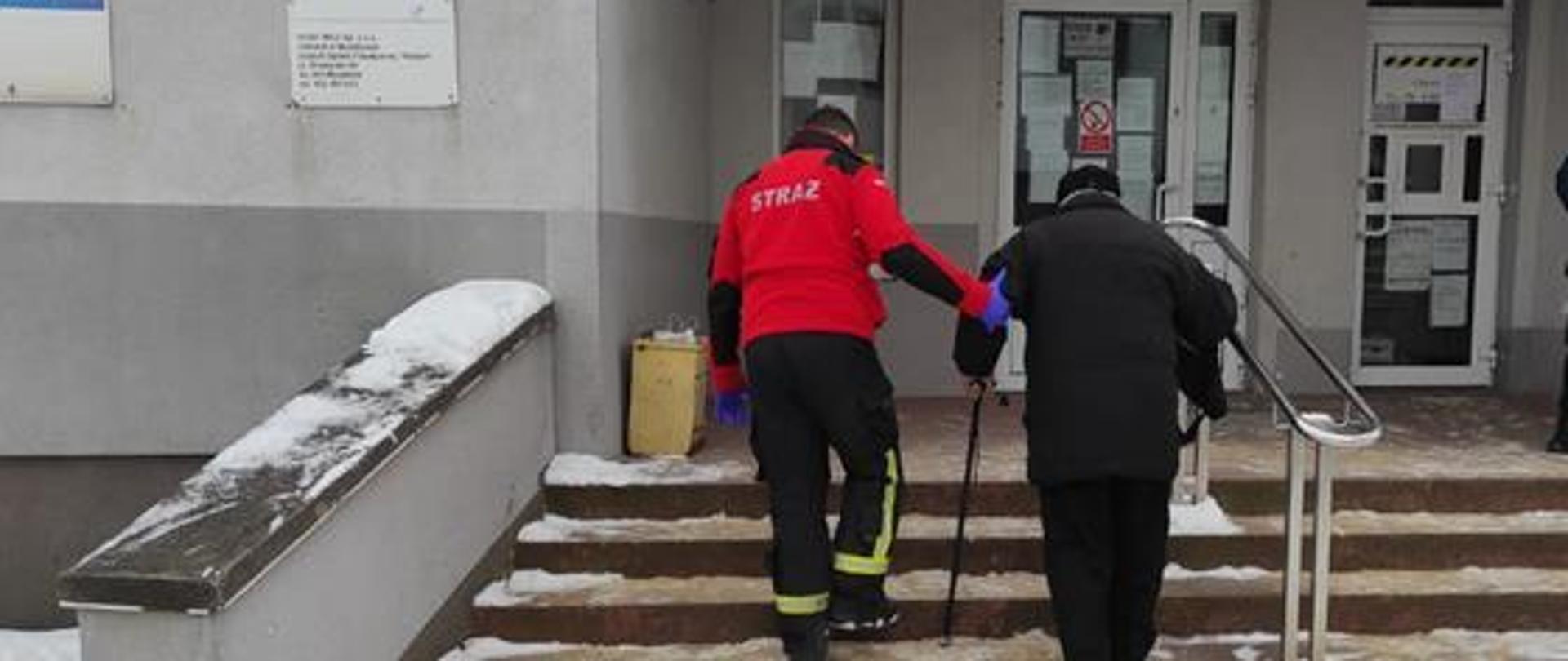 Fotografia przedstawia strażaka prowadzącego osobę starszą po schodach. Widok z tyłu. 