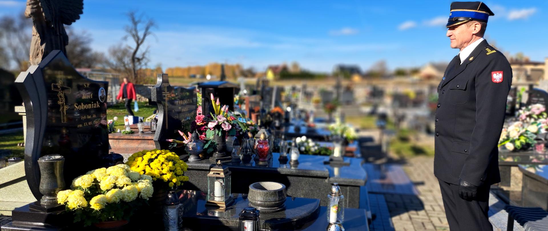 Alejka cmentarna, funkcjonariusz CS PSP w mundurze galowym stoi w zadumie przed nagrobkiem