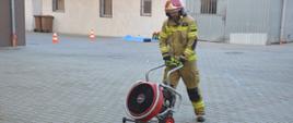 Ćwiczenia w Zakładzie Karnym w Rawiczu. Zdjęcie przedstawia tzw." teren ścisły". Na tle budynków znajduje się strażak w ubraniu specjalnym i w czerwonym hełmie, który pcha agregat oddymiający. Z lewej strony, w oddali stoja dwa biało-czerwone pachołki. 