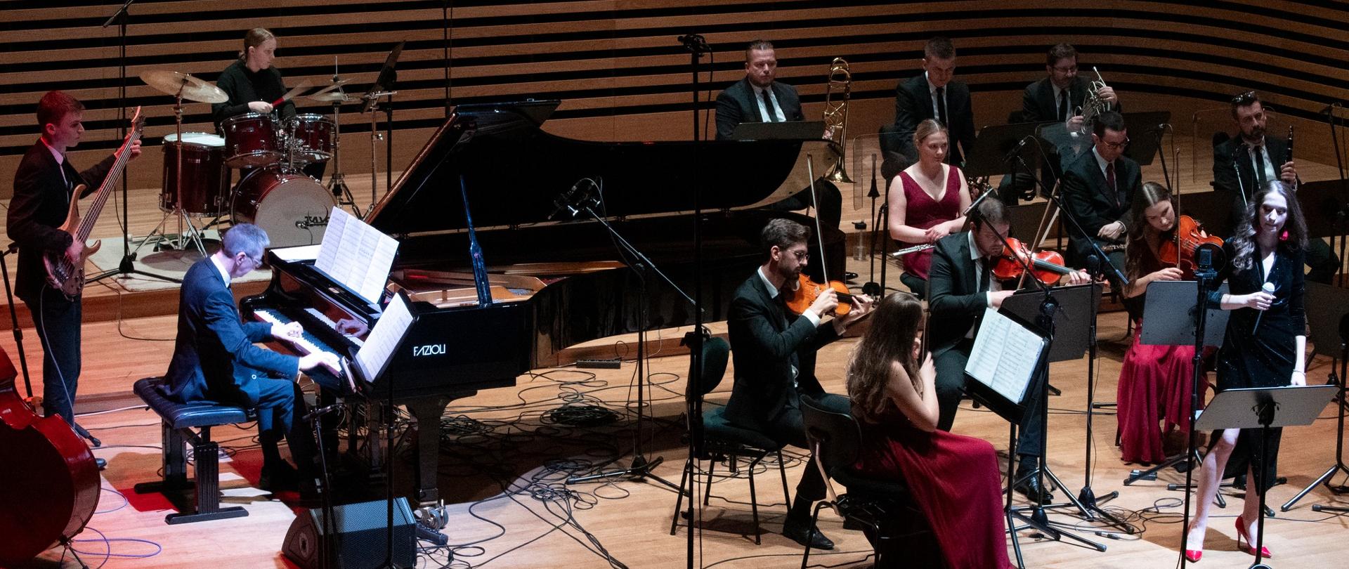 Muzycy zespołu grają na estradzie, po prawej stronie przed nimi stoi kobieta z mikrofonem.