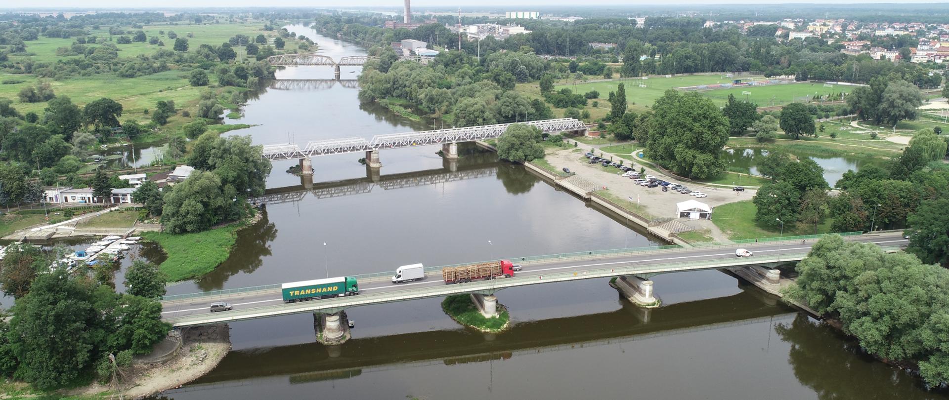 Zdjęcie z powietrza przedstawiające rzekę Wartę oraz rozpięte nad nią trzy mosty drogowe i kolejowe w otoczeniu nadrzecznej zieleni i zabudowy miejskiej.