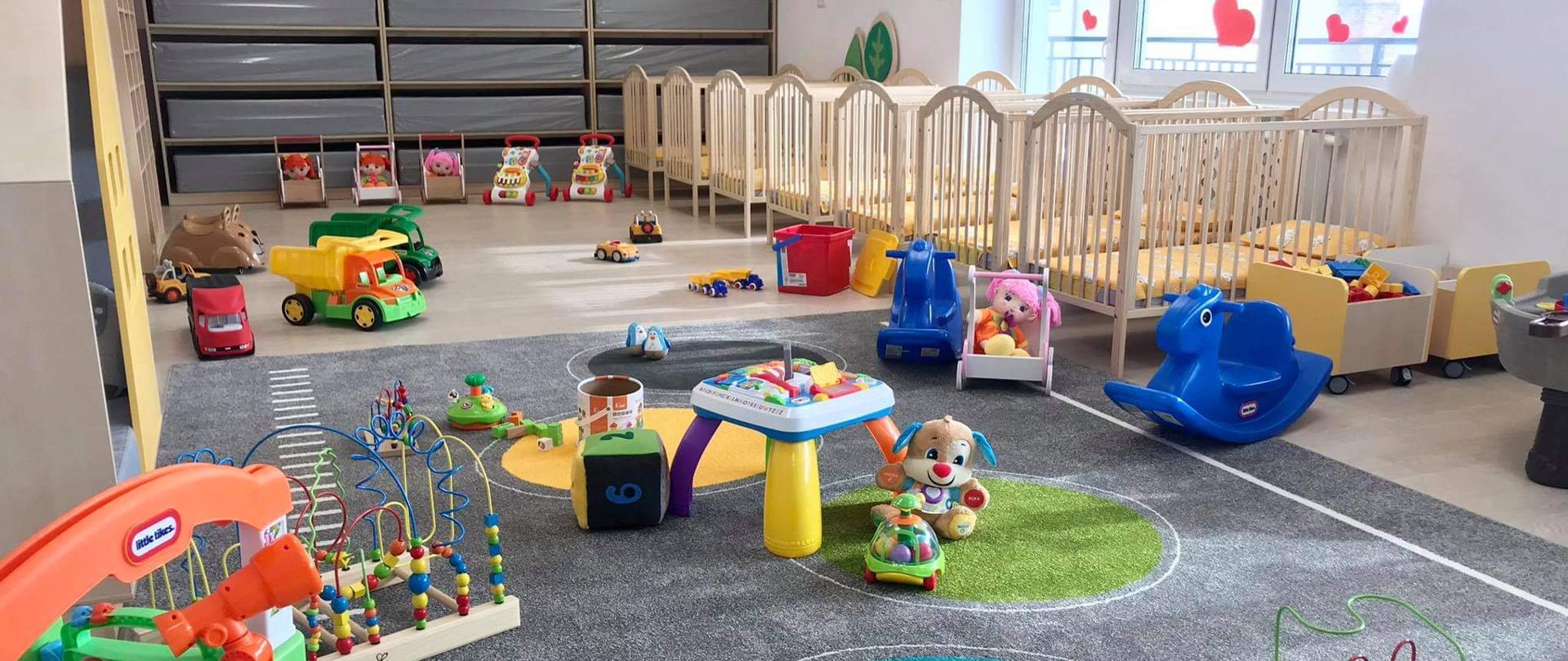 Uroczyste otwarcie żłobka w Gorzowie Śląskim. Na zdjęciu jedna z sal zabaw dla dzieci. Na pierwszym planie kolorowy dywan oraz ustawione na nim zabawki sensoryczne. W tle ustawione rzędem 7 łóżek dziecięcych. 