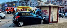 Na pierwszym planie widoczny jest czarny samochód osobowy (marki Hyundai), który z dużą siłą uderzył w niewielki kiosk z napisem „TAXI”. Przednia część auta jest wbita w konstrukcję budki, a pokrywa silnika została wygięta. Kiosk, pokryty graffiti, uległ znacznym uszkodzeniom, a jego drzwi są lekko uchylone.
W tle, za uszkodzonym pojazdem, pracują służby ratownicze:
Grupa strażaków w pełnym umundurowaniu bojowym (hełmy, kurtki typu nomex) zabezpiecza miejsce zdarzenia.
Widoczny jest wóz strażacki (ciężarowy samochód ratowniczo-gaśniczy) oraz żółta karetka pogotowia z włączoną sygnalizacją świetlną.
W oddali widać zabudowę miejską, kamienice oraz znaki drogowe (m.in. informację o parkingu płatnym).