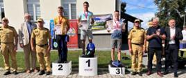 Na zdjęciu widzimy strażaków na podium pozujących do zdjęcia z pucharami i dyplomami, obok stoją komendanci w ubraniach służbowych oraz lokalne władze. 