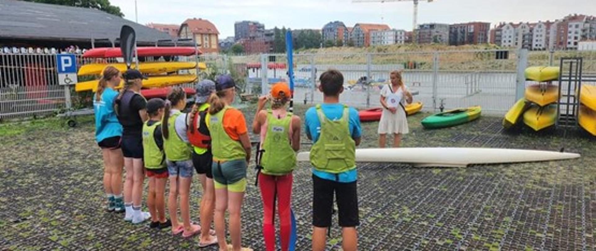 Kobieta w kremowej sukience z teczką z logo Powiatowej Stacji Sanitarno-Epidemiologicznej w Gdańsku stoi zwrócona w stronę dzieci w kamizelkach ratunkowych. Na stojakach znajdują się ułożone kajaki.
