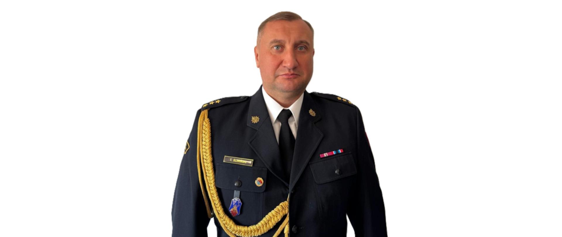 Zdjęcie przedstawia mężczyznę w formalnym mundurze służbowym. Ma krótkie, jasne włosy i poważny wyraz twarzy. Mundur jest granatowy, z krawatem, ozdobnymi złotymi sznurami na ramieniu oraz odznaczeniami i naszywkami na piersi. Tło jest jasne i jednolite.