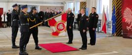 Komendant Wojewódzki, odchodzący Komendant Powiatowy i nowy Komendant Powiatowy stoją w garażu Komendy Powiatowej w Lubaniu podczas uroczystości zdania i przyjęcia obowiązków Komendanta Powiatowego Państwowej Straży Pożarnej w Lubaniu. Po lewej stronie poczet sztandarowy ze sztandarem Komendy Powiatowej Państwowej Straży Pożarnej w Lubaniu. W tle stoją funkcjonariusze i osoby cywilne, strażacy są w mundurach wyjściowych. Po prawej stronie flagi Polski, Unii Europejskiej oraz flaga z logiem Państwowej Straży Pożarnej.