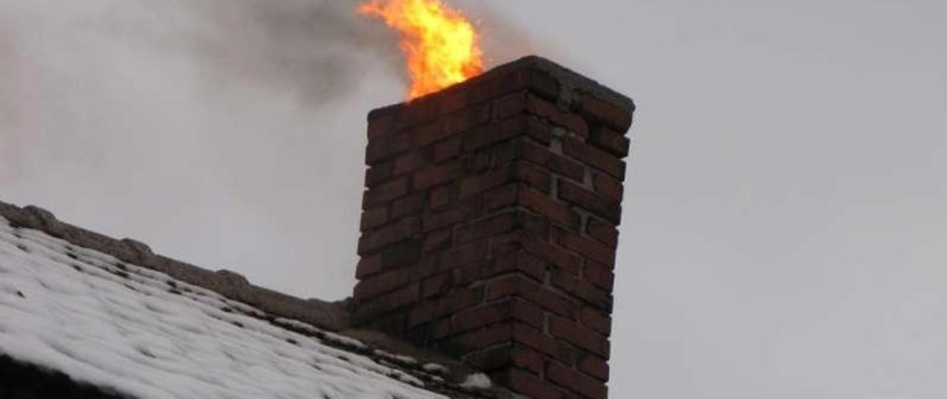 Zdjęcie przedstawia komin na dachu budynku, z którego wydobywa się ogień płonącej sadzy.