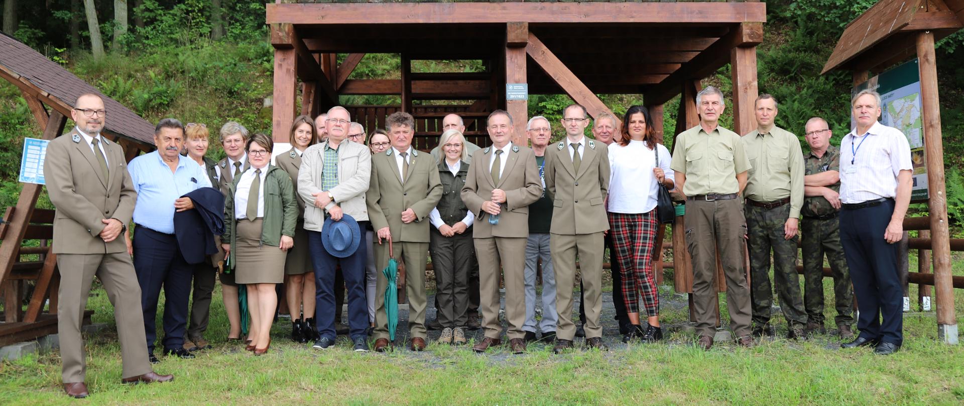 Grupa składająca się z kilkunastu osób (kobiety i mężczyźni), pozująca na tle drewnianej wiaty widokowej. Po prawej stronie widać fragment tablicy informacyjnej, a po lewej kawałek zadaszonej drewnianej wiaty,