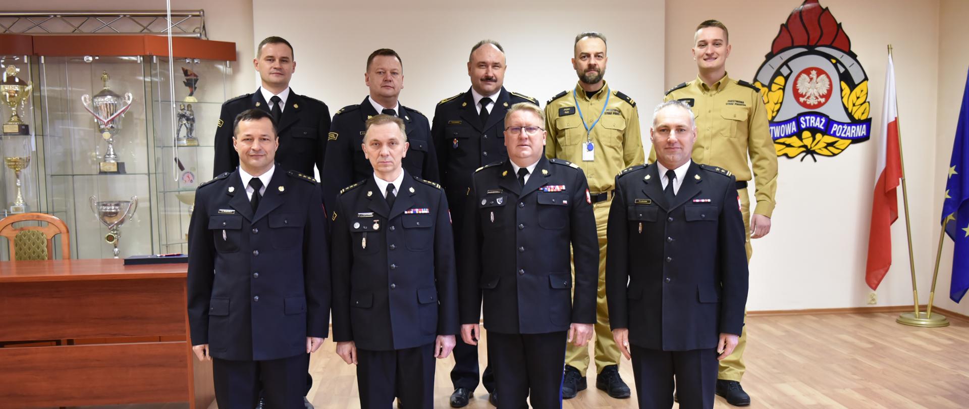 9 strażaków stojących w dwóch rzędach, drugi rząd stoi na podwyższeniu, pozują do zdjęcia w sali konferencyjnej