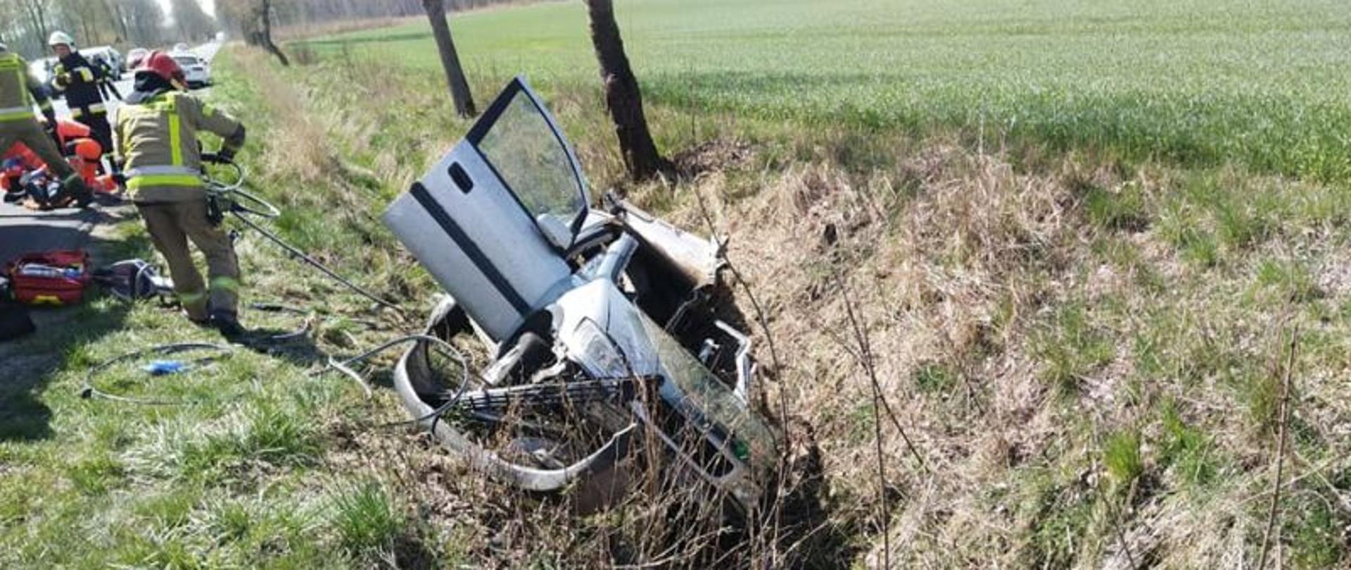 Zdjęcie wykonane w dzień. Przedstawia auto leżące w rowie, na boku od strony kierowcy. Widać przód auta. Auto koloru srebrnego. Ma odgięty i wycięty dach. W tle strażacy i wóz gaśniczy, Auta stoją wzdłuż drogi.