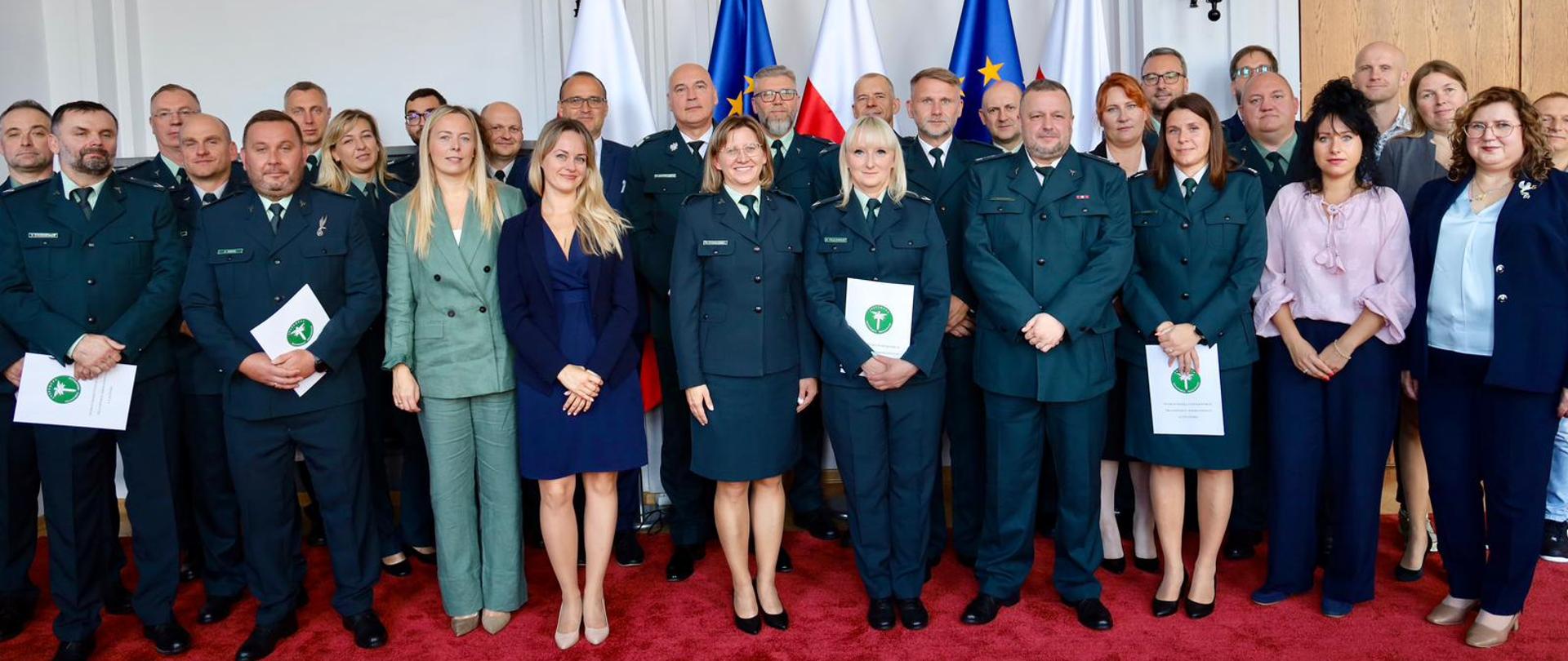 Grupa kilkudziesięciu osób, w tym funkcjonariusze w ciemnozielonych mundurach oraz osoby w strojach cywilnych, pozuje do pamiątkowego zdjęcia. W tle widoczne są flagi Polski i Unii Europejskiej oraz zawieszone na ścianie elementy dekoracyjne. Niektóre osoby w pierwszych rzędach trzymają w dłoniach teczki.