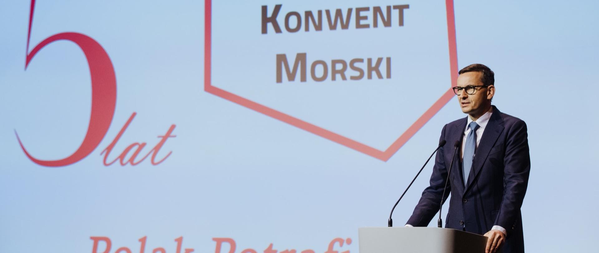 Premier Mateusz Morawiecki podczas wypowiedzi na Konwencie Morskim w Gdańsku. Szef rządu stoi przy mównicy z mikrofonami. W tle jest wyświetlana grafika: 5 lat Konwentu Morskiego z hasłem Polak Potrafi.