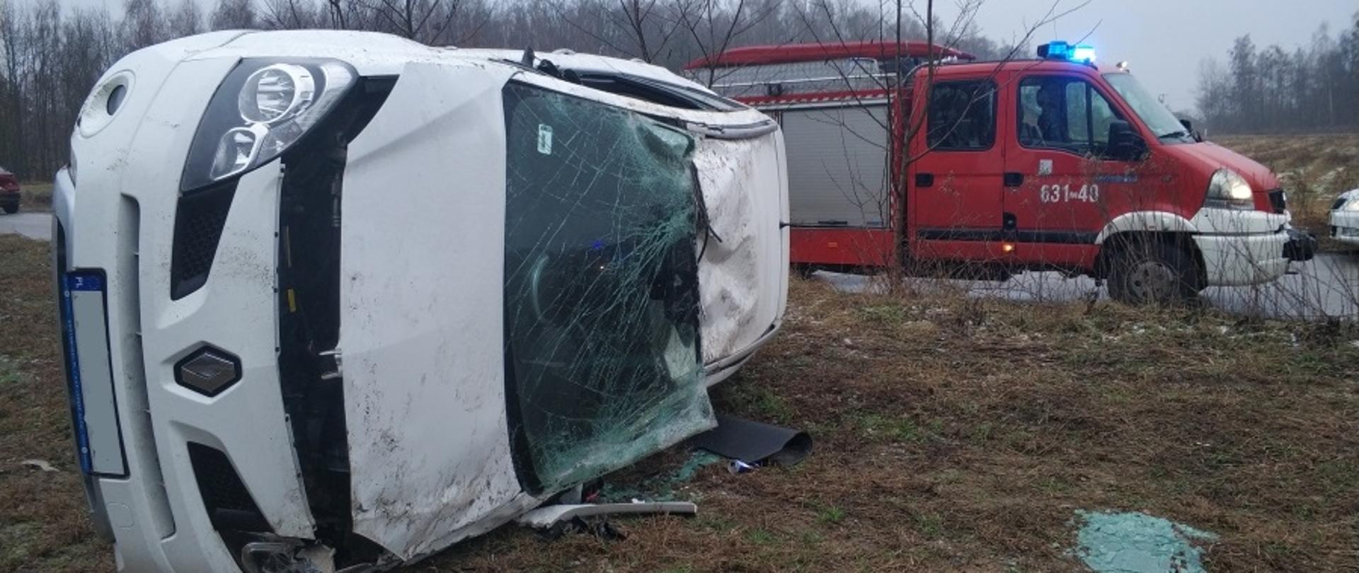 Na zdjęciu widoczny samochód osobowy koloru białego leżący poza jezdnią, na poboczu, na lewym boku. Widać rozbitą przednią szybę, uszkodzony dach. Na drugim planie samochód straży pożarnej.