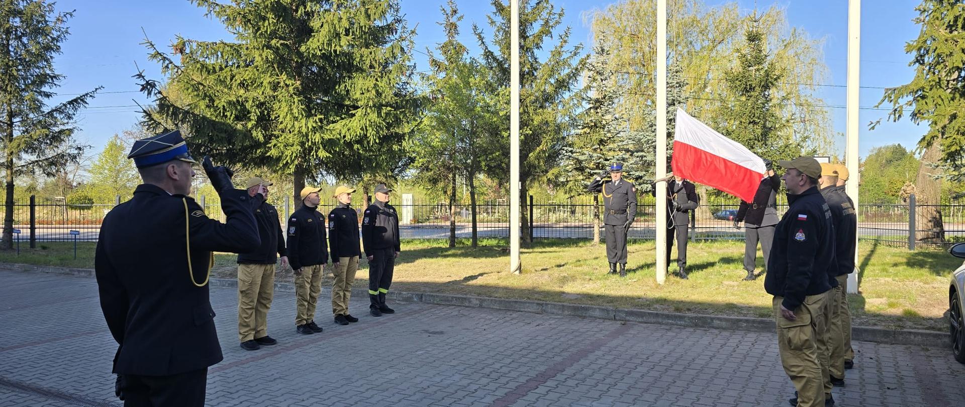 Na zdjęciu widoczna jest uroczystość podniesienia flagi państwowej Rzeczypospolitej Polskiej, która odbywa się na zewnątrz.
Główne elementy i szczegóły:
Uczestnicy: W ceremonii bierze udział grupa osób w mundurach, przypominających umundurowanie straży pożarnej lub innej służby mundurowej.
Główny moment: Na maszcie podnoszona jest biało-czerwona flaga.
Postacie i ich role:
Po lewej stronie: Mężczyzna w ciemnym mundurze galowym stoi tyłem i oddaje honory (salutuje).
Przed nim: Stoi grupa mężczyzn w ciemnych polarach/bluzach i beżowych spodniach, ustawiona w szeregu na baczność.
Przy maszcie: Jedna osoba podnosi flagę, a druga stoi obok niej.
Po prawej stronie: Kolejni uczestnicy w podobnych mundurach obserwują ceremonię.
Otoczenie: Wydarzenie odbywa się na placu wyłożonym kostką brukową. W tle widoczny jest trawnik, drzewa (głównie iglaste), ogrodzenie oraz bezchmurne, niebieskie niebo, co wskazuje na słoneczną pogodę.
Całość ma charakter oficjalny i patriotyczny.