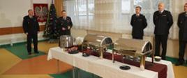 Widok na strażacką świetlicę. W rogu pomieszczenia choinka oraz Polskie godło. Na środku pomieszczenia przygotowana wigilia. Jedzenie stoi na stole. Za stołem bliżej choinki Komendant oraz jego Zastępca w mundurach galowych. Po prawej stronie wzdłuż ściany kolei strażacy odświętnie ubrani