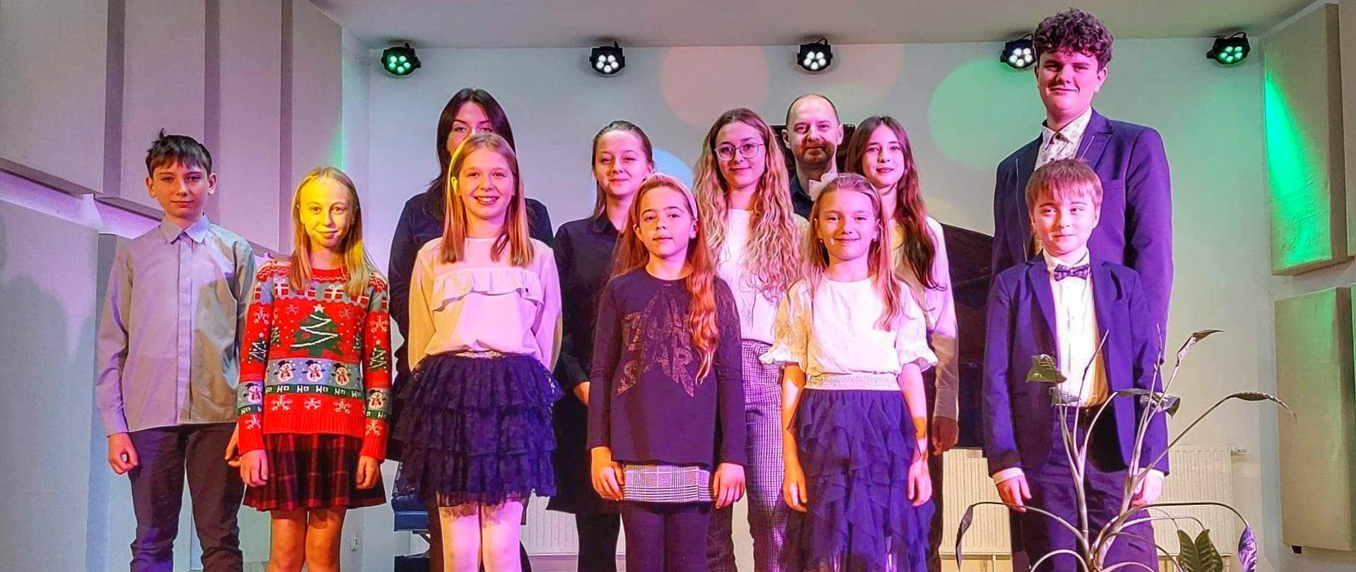 Zdjęcie przedstawia uśmiechniętych uczniów klasy fortepianiu stojących na scenie szkoły wraz z nauczycielem. Za uczniami na środku sceny stoi czarny fortepian, a przed nimi po prawej stronie 3 białe donice z kwiatami. 