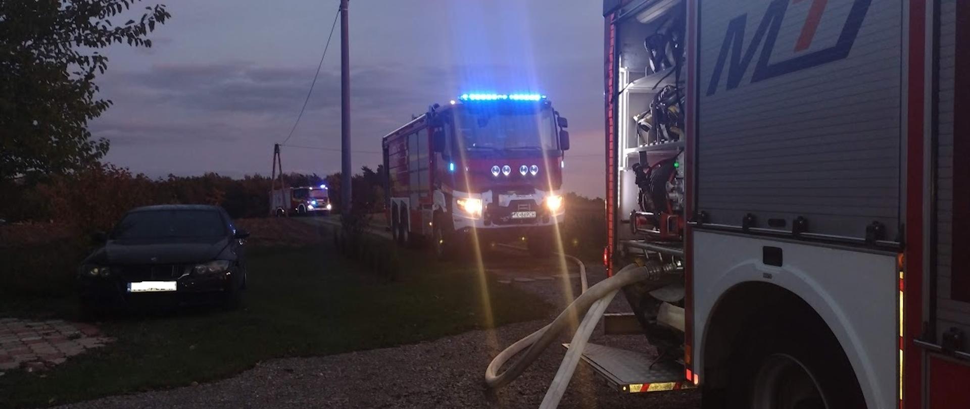 Na zdjeciu widać 3 samochody gaśnicze podczas akcji gaszenia pożaru stodoły i grażu. Na pierwszym planie widać węże pożarnicze podłączone do dwóch strażackich samochodów. Zdjecie wykonane w porze wieczornej. 