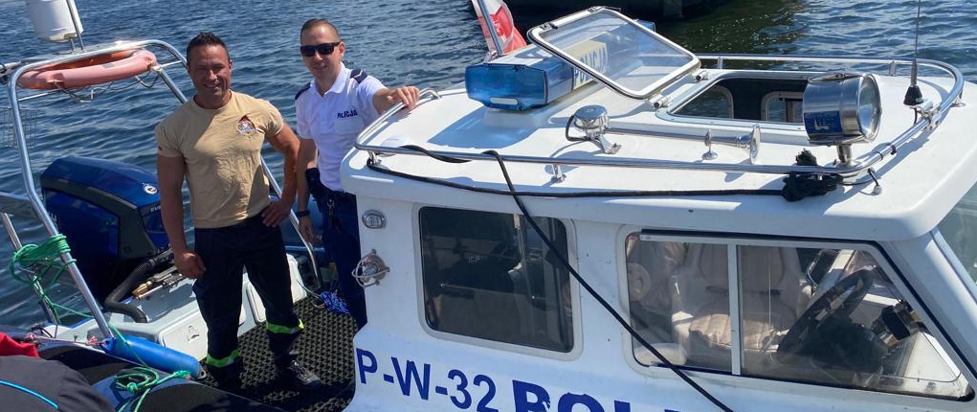 Na zdjęciu widoczna niebiesko-biała prawa burta łodzi z napisem P W 32 POLICJA. Na łodzi stoi dwóch uśmiechniętych mężczyzn. Pierwszy z lewej w piaskowej koszulce i ciemnych spodniach z jasnozieloną taśmą pod kolanami i czarnych butach, drugi w okularach, białej koszulce z napisem na piersi POLICJA i ciemnych spodniach, trzyma rękę na relingu dachu. W tle otwarta woda zalewu, w oddali ledwie widoczna zieleń drzew i błękitne niebo z białymi obłokami. Tuż za łodzią kadłub betonowca.