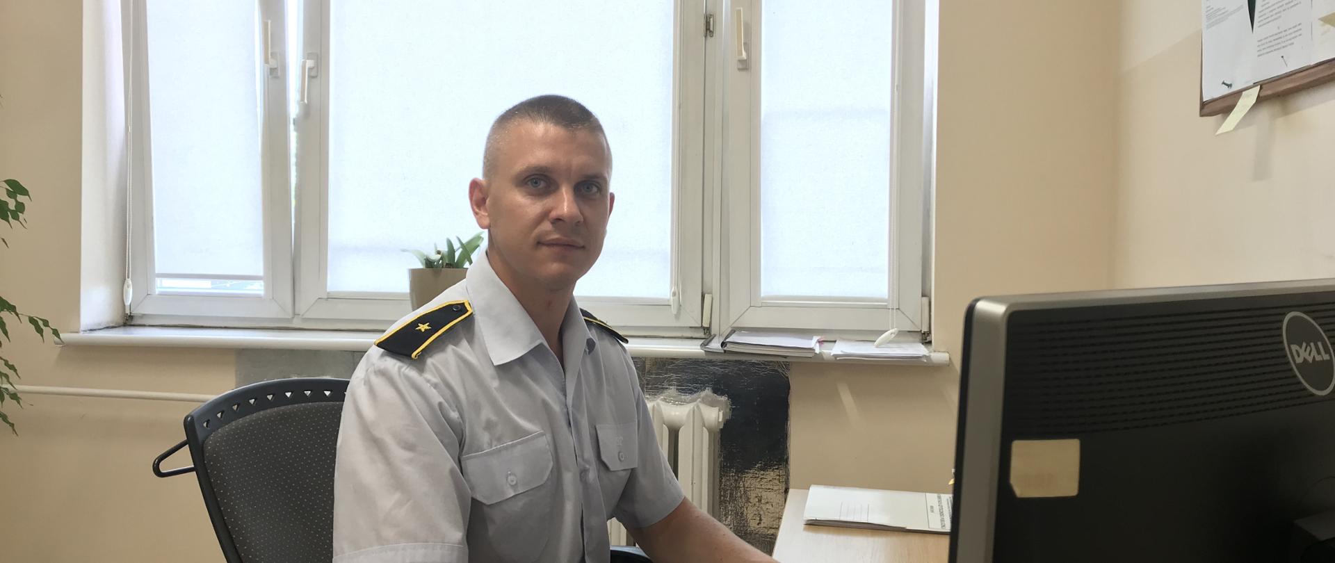 Zdjęcie zrobione w biurze, funkcjonariusz siedzi przed biurkiem z rękami ułożonymi na blacie. Strażak ubrany w niebieską koszule z pagonami.