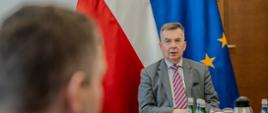 W sali przy podłużnym stole siedzą ludzie, u szczytu stołu minister Wieczorek mówi, za nim pod ścianą flagi Polski i UE.