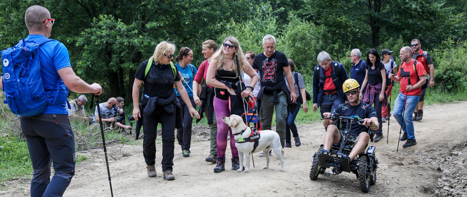 Grupa osób na szlaku górskim, w środku kobieta z psem przewodnikiem, po prawej na specjalnym wózku elektrycznym Krystian Herba