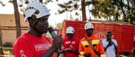 Uczestnik szkolenia w białym kasku i czerwonej koszulce z napisem „Firefighter” przemawia przez mikrofon. W tle stoją trzy osoby, w tym jedna w pomarańczowym kombinezonie ochronnym i białym kasku. Za grupą znajduje się czerwony pojazd techniczny.