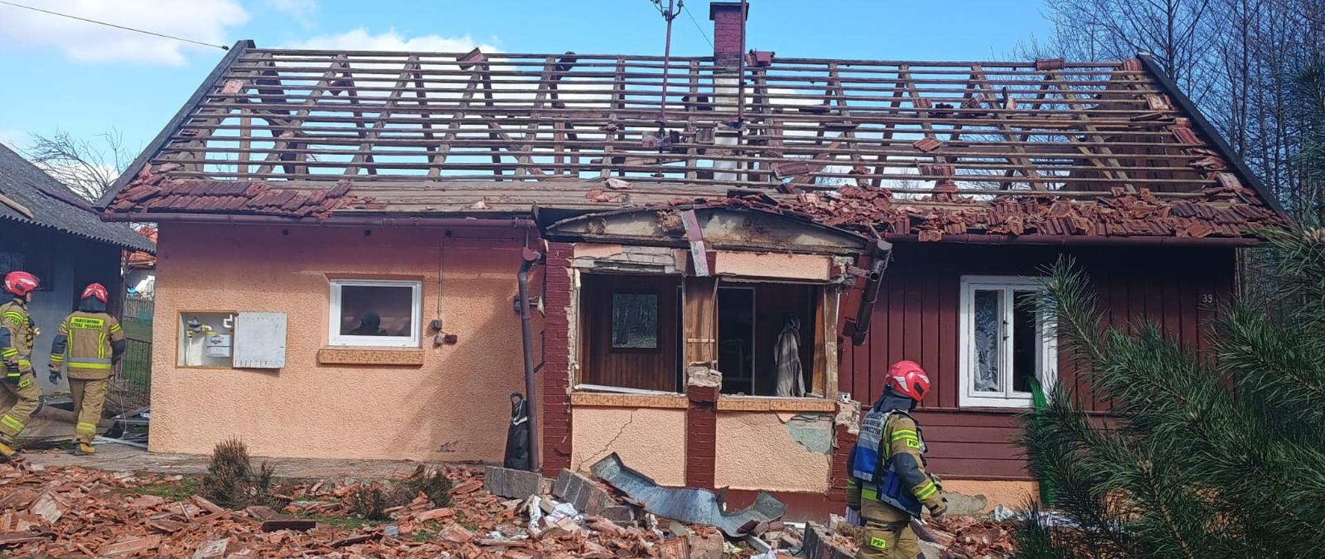 Dom z uszkodzonym dachem i gankiem, po lewej stronie strażak w ubraniu specjalnym, w tle z lewej strony 2 strażaków i budynek gospodarczy
