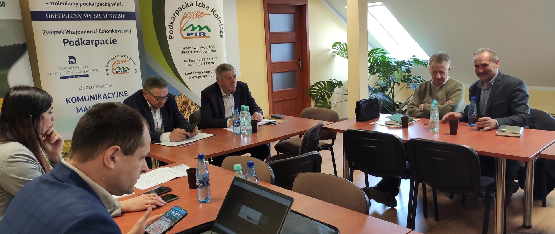 Grupa osób siedząca w sali przy stołach. Mężczyzna na pierwszym planie robi coś na laptopie. W tle widać banery z logiem instytucji. W tle stoi też zielony kwiat. Pomieszczenie jest jasne.