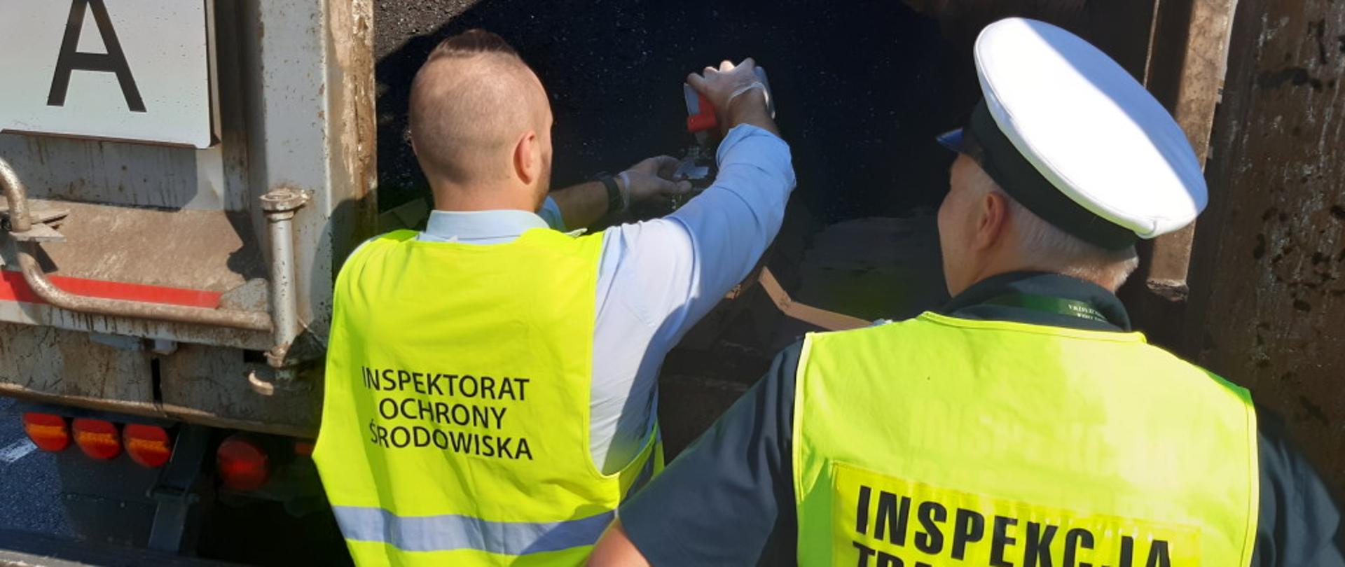 Dwóch mężczyzn w odblaskowych kamizelkach. Za nimi otwarta naczepa samochodu ciężarowego na której znajduje się odpad w formie sypkiej. 
