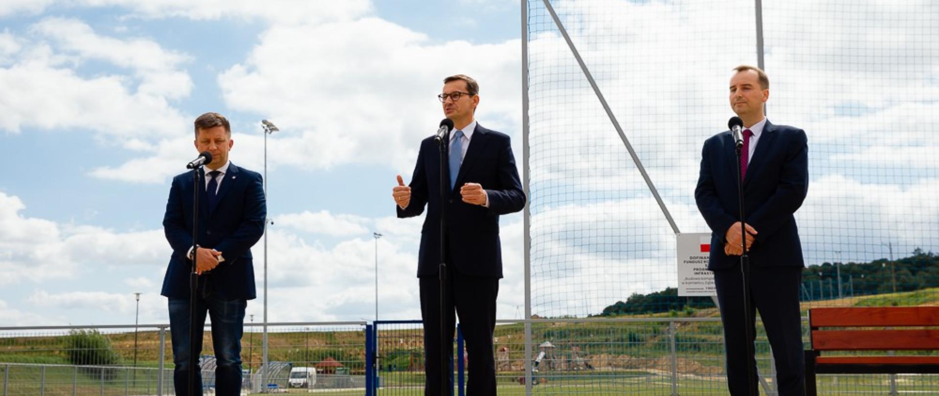 Konferencja prasowa na tle boiska piłkarskiego.