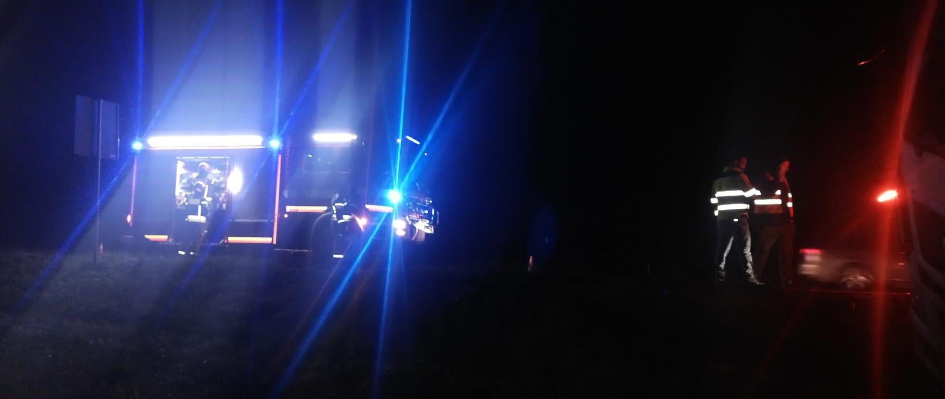 Zdjęcie zrobione nocą. Na drodze ustawione suto strażackie, oświetlają teren, włączone niebieskie światła ostrzegawcze. Z prawej strony stoją dwie osoby.