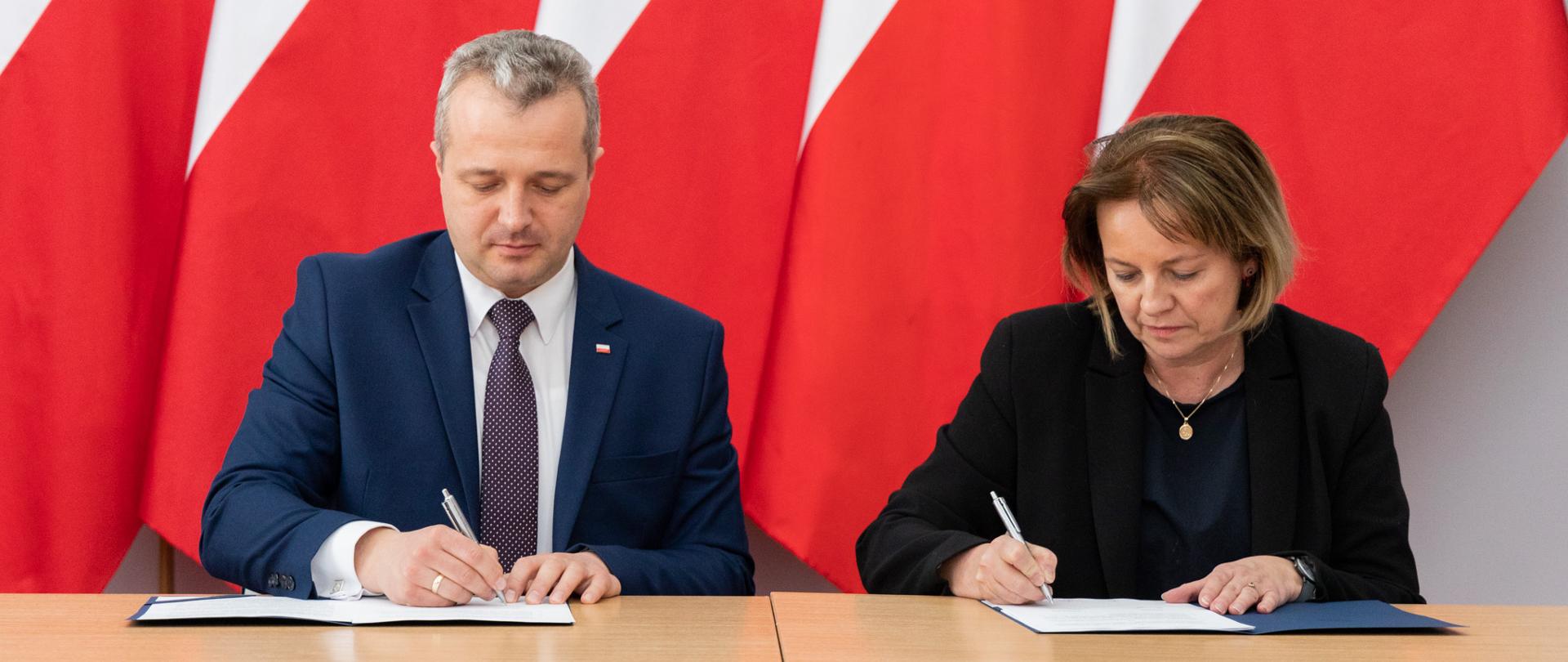 Dwie osoby siedzą i podpisują umowy. 