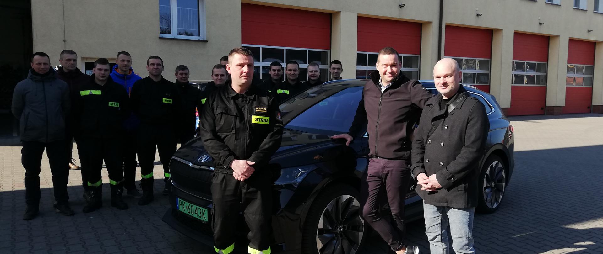 Zdjęcie przedstawia grupę osób stojących przy samochodzie z napędem elektrycznym koloru czarnego na tle budynku jednostki ratowniczo-gaśniczej.