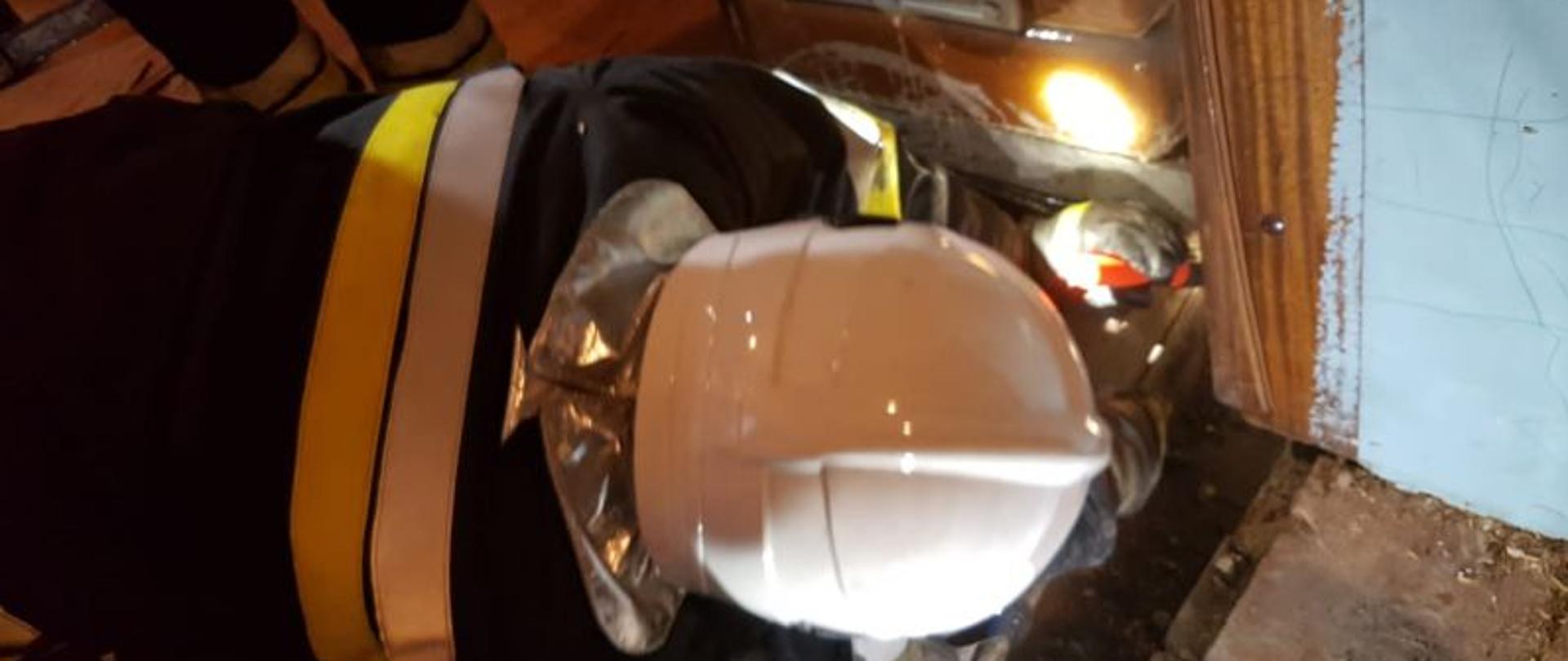 Na zdjęciu strażak w białym hełmie. Strażak pochylony nad piecem. Strażak świeci latarką pod kaflowy brązowy piec w poszukiwaniu zarzewi ognia