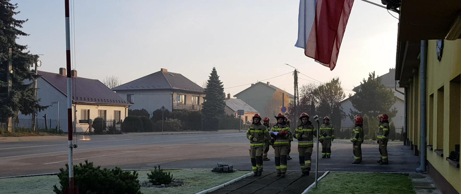 trzech strażaków maszeruje z flagą, w tle strażacy stoją w dwuszeregi.