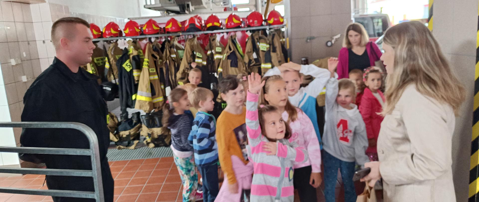 Garaż jednostki ratowniczo-gaśniczej, grupa dzieci wraz z opiekunkami w czasie pogadanki na temat służby w Państwowej Straży Pożarnej prowadzonej przez strażaka.